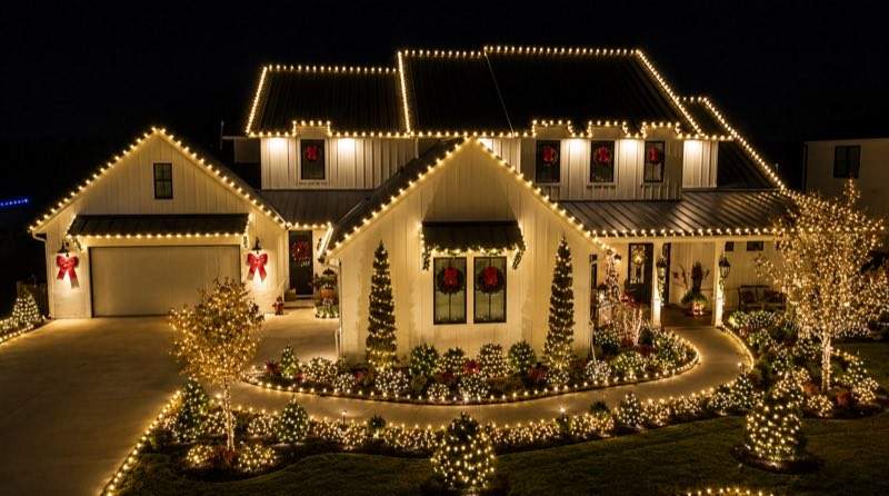 White farmhouse fully outlined in warm white lights with lit wreaths, bows, and a landscaped light display